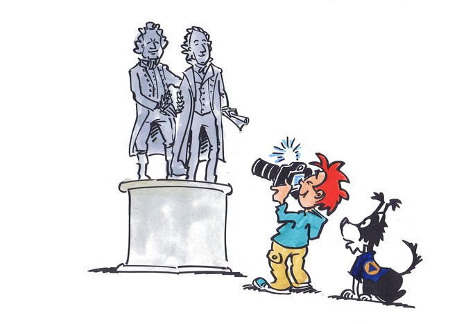Max und Flocke stehen vor einem Denkmal mit einer Goethe und einer Schiller Statue. Max fotografiert das Denkmal.